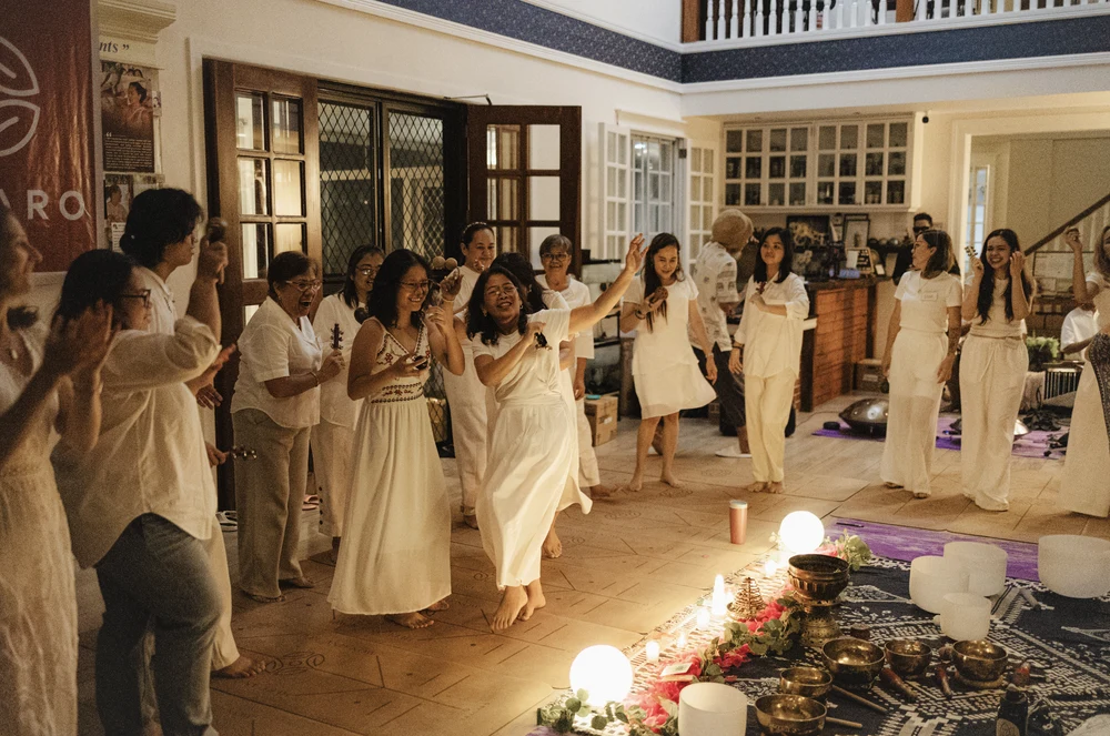 Women in white dancing joyfully in a circle around a crystal sound-bowl setup indoors at Tallaro